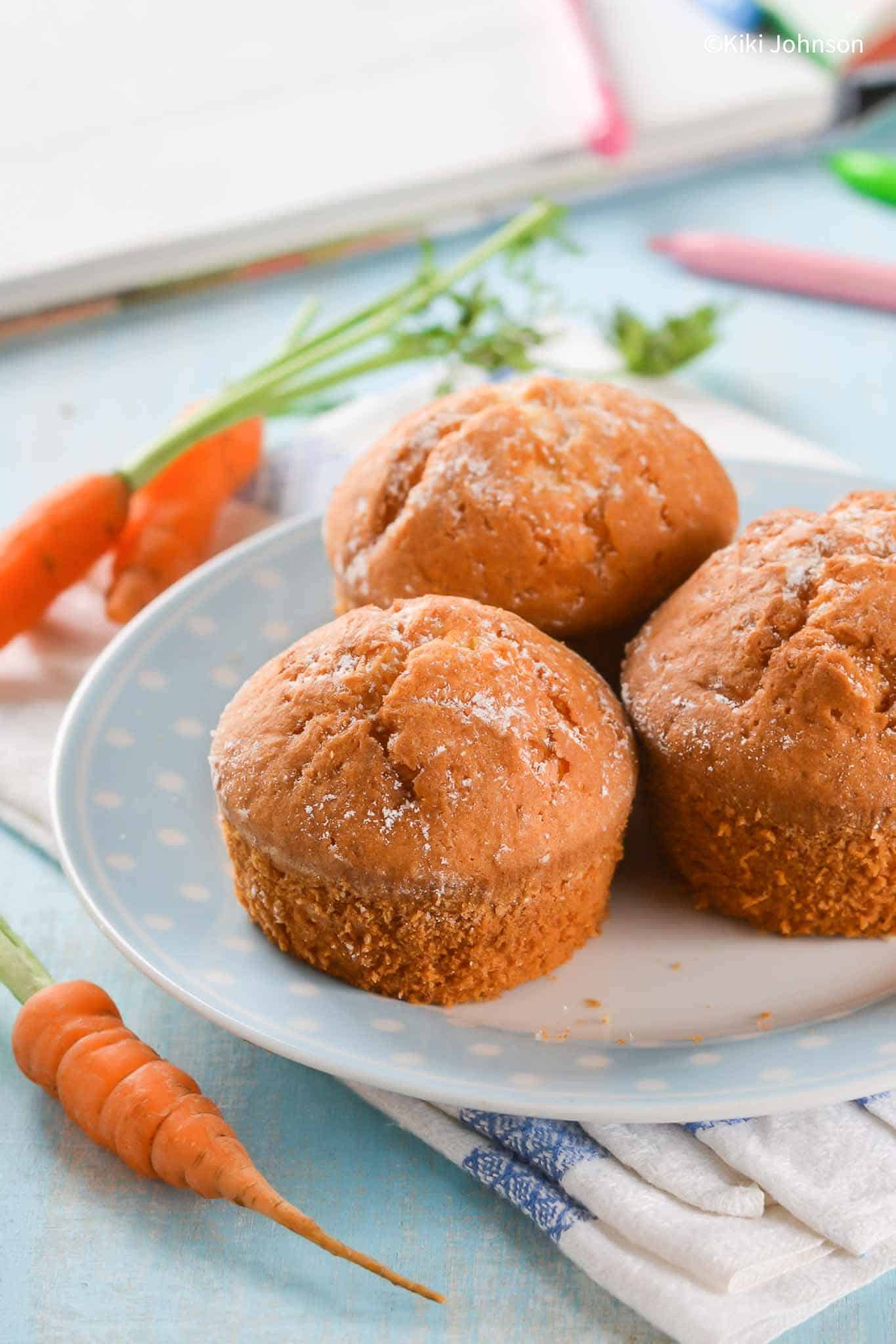 three carrot banana muffins on a blue plate with fresh carrots surrounding the plate