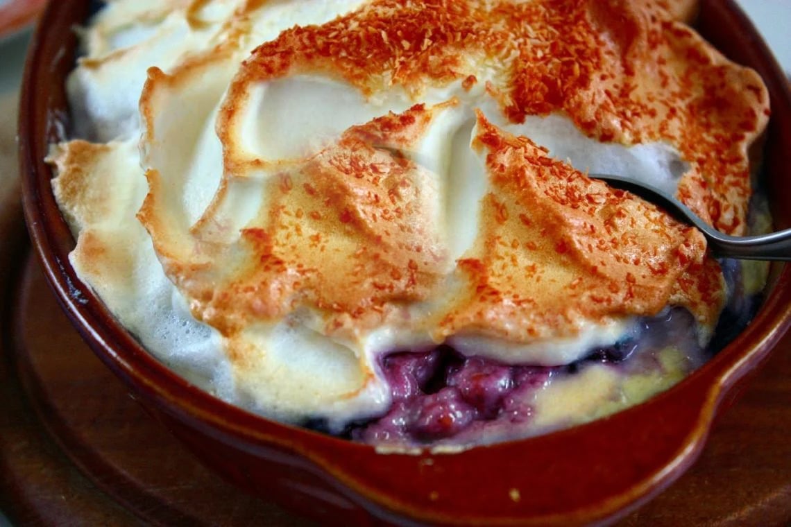 a spoon digging into a casserole dish with blueberry baked rice pudding topped with golden meringue topping 