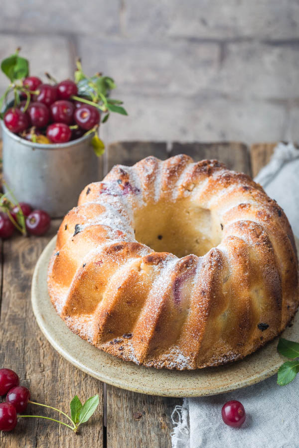 a white chocolate and fresh cherry bundt cake on a cake platter with a metal cup of cherries in the background
