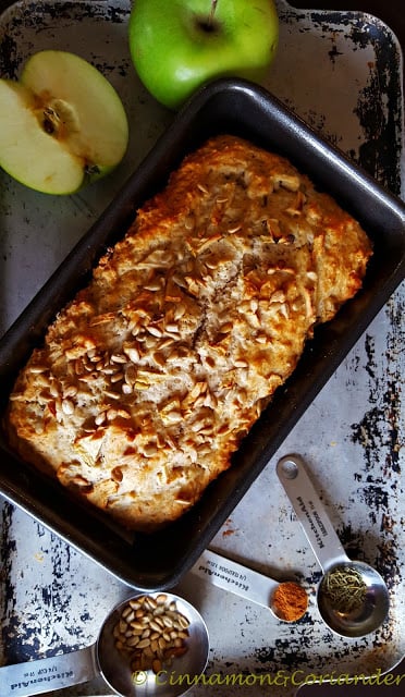 ein saftiges Apfelbrot in der Kastenform mit Sonnenblumenkernen bestreut auf einem silbernen Blech mit frischen Äpfeln im Hintergrund.