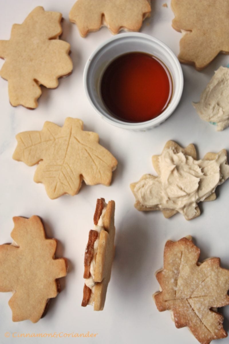 maple leaf sandwich cookies whimsically arranged on a marble countertop with a small dish of maple syrup on the side