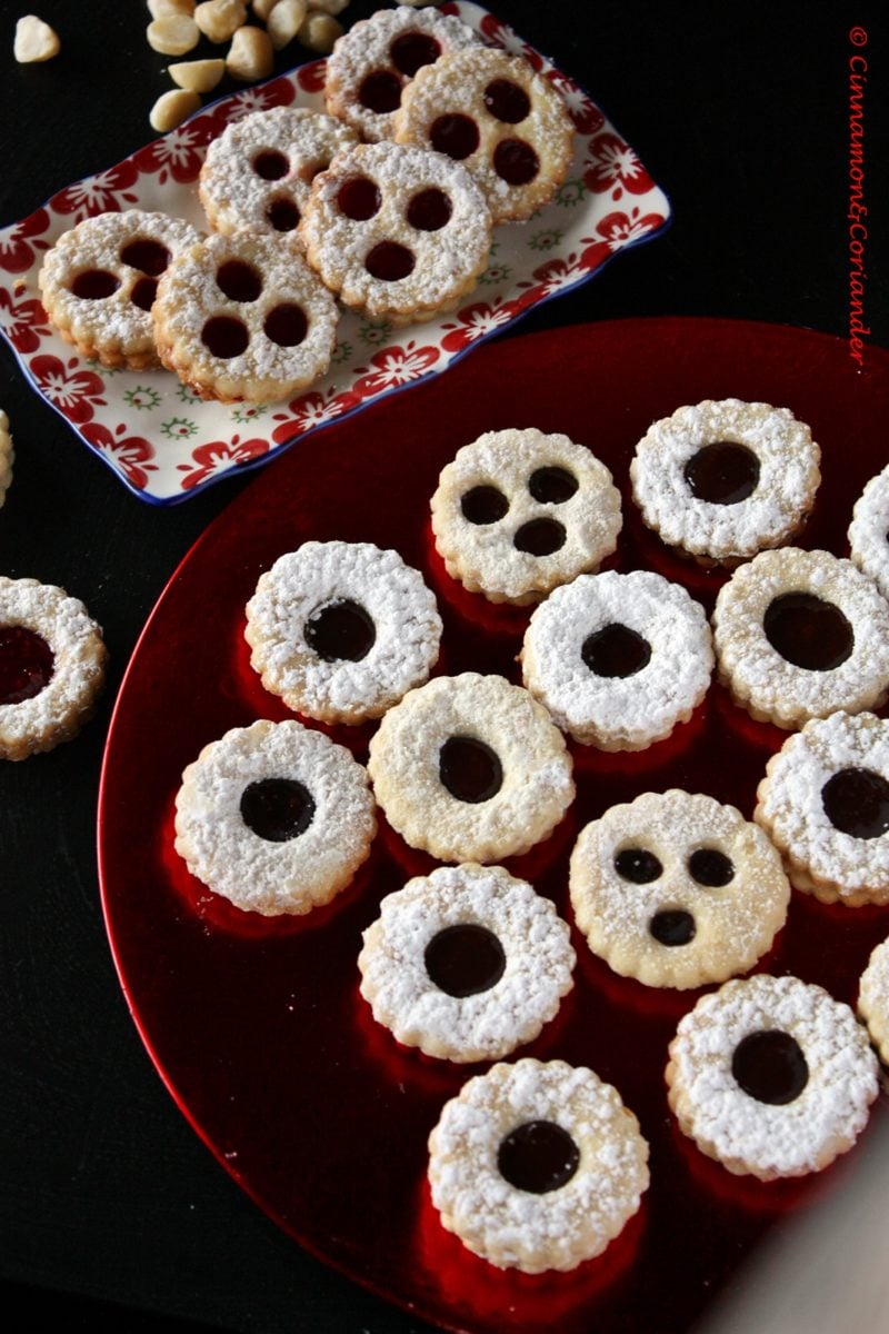 a variety of German Christmas cookies like Macadamia Cherry Jam Sandwich Cookies called Spitzbuben and Black Forest Sandwich Cookies arranged on a red plate