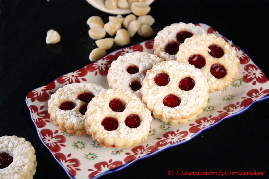 German Christmas Cookies (Spitzbuben) filled with Cherry Jam on a small plate with some macadamia nuts in the background