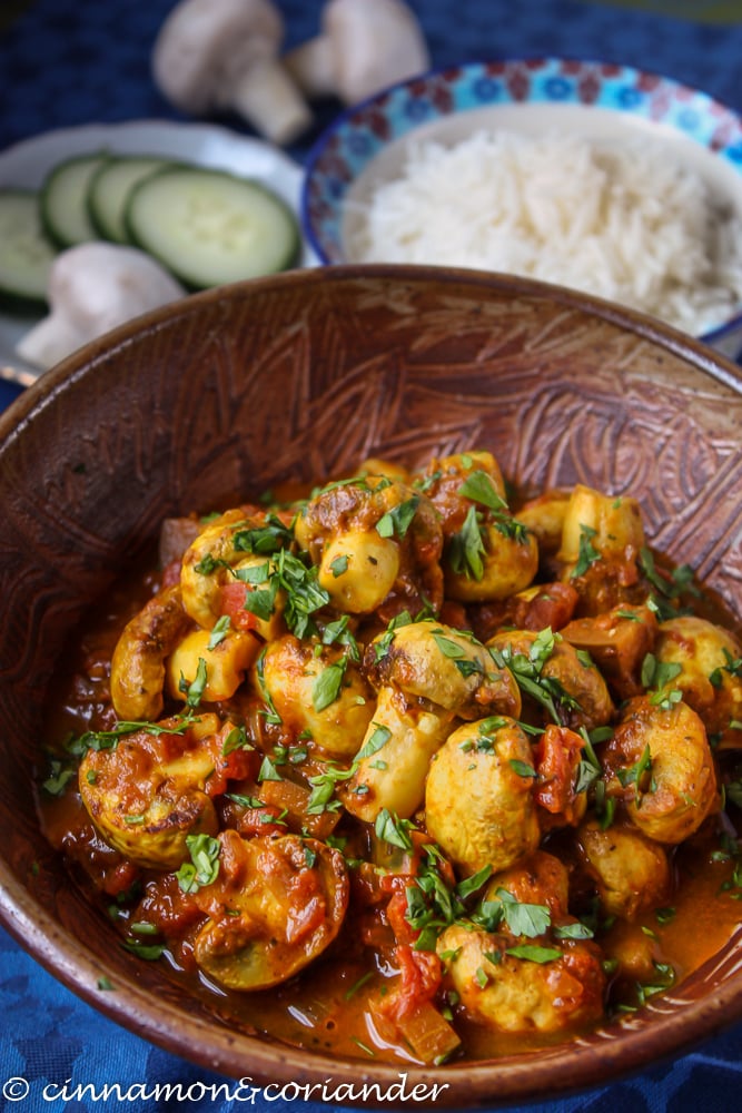 Vegetarian Mushroom Tikka Masala served in a rustic Indian bowl with a dish of rice and some slice veggies in the background