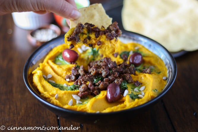 a chip being dipped into Loaded Kabocha Squash Hummus with ground lamb and lemon parsley sauce