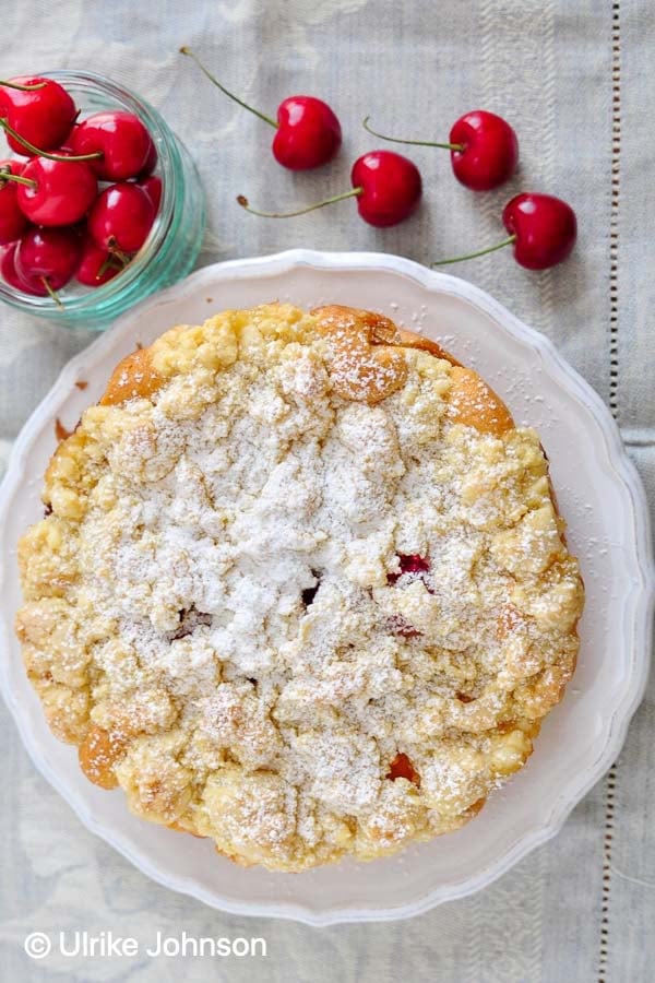 Omas Aprikosen Streuselkuchen mit Kirschen und mit Puderzucker bestreuten Butterstreuseln auf einem Tortenteller