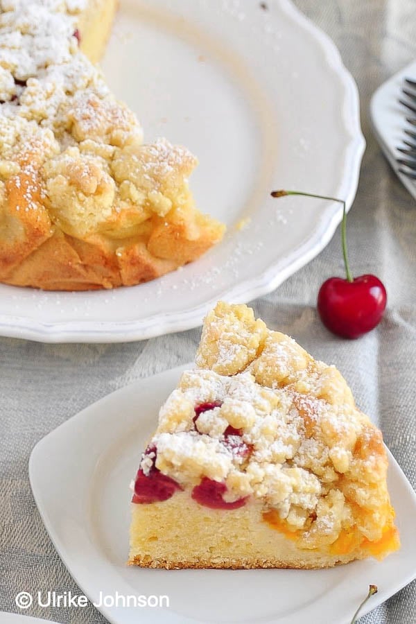 ein Kuchenteller mit einem Stück Aprikosen Streuselkuchen mit Kirschen neben einem angeschnittenen Obstkuchen