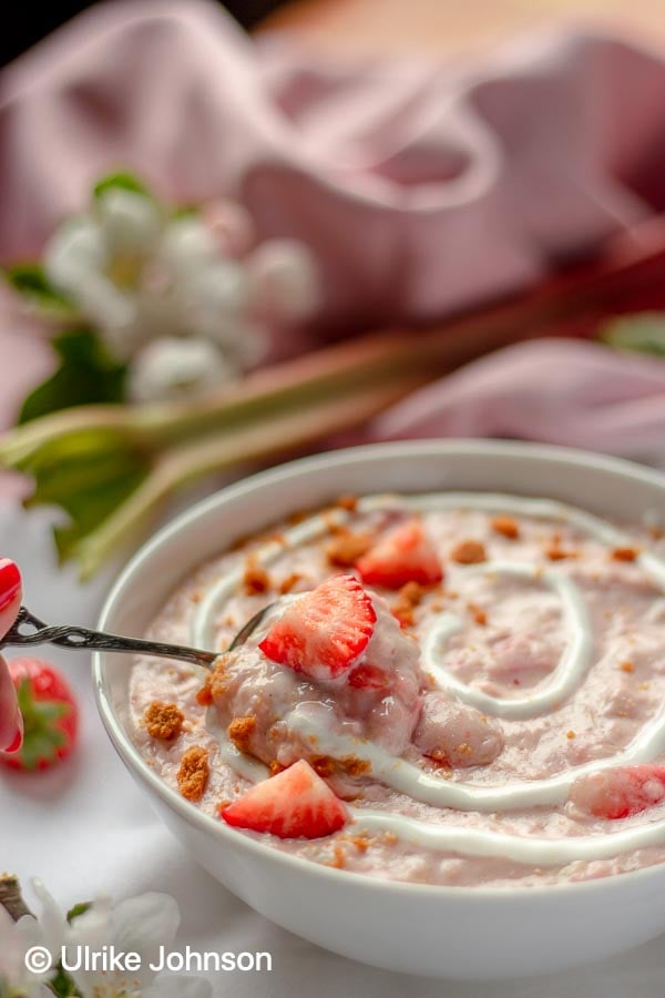 a spoon being dipped into an oatmeal bowl with rhubarb strawberry cheesecake porridge decorated with fresh strawberries and cookie crumbs 