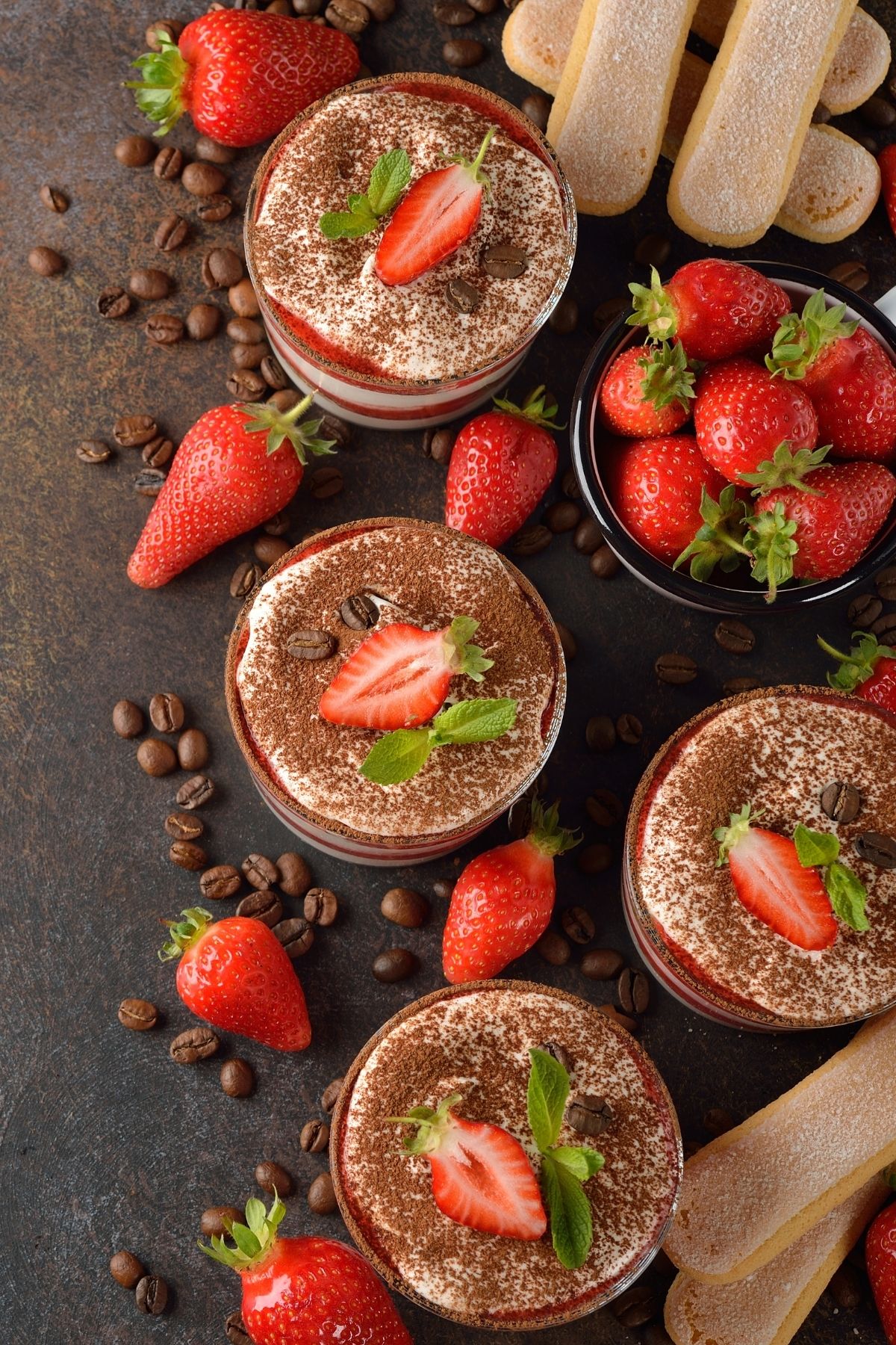 overhead shot of 4 dessert glasses with strawberry tiramisu surrounded with ladyfingers and coffee beans