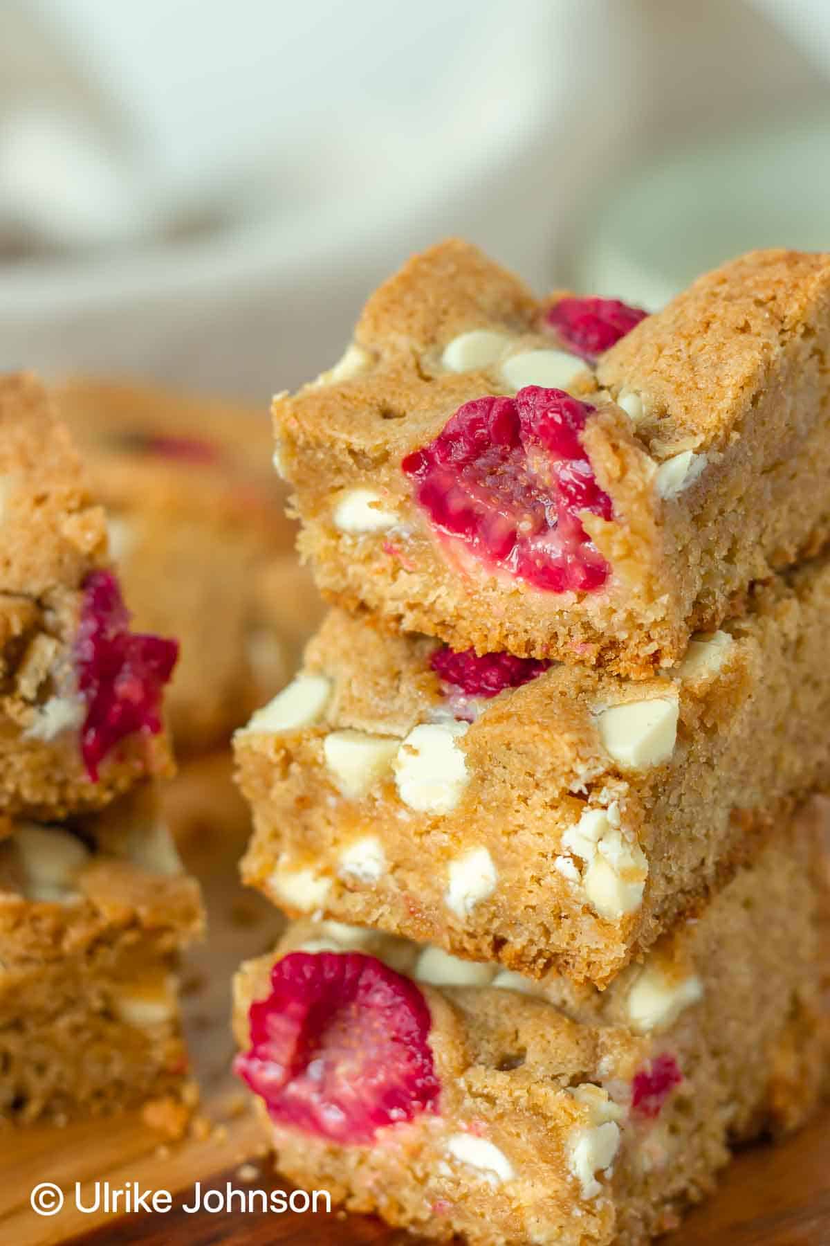 chewy glutenfree raspberry and white choc blondies stacked on a rustic wooden board