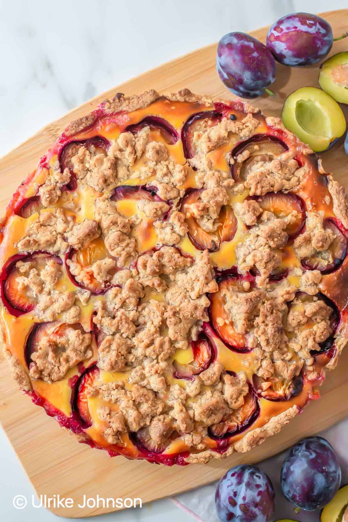 German Plum Custard Tart with streusel topping on a wooden board surrounded by Italian prune plumbs