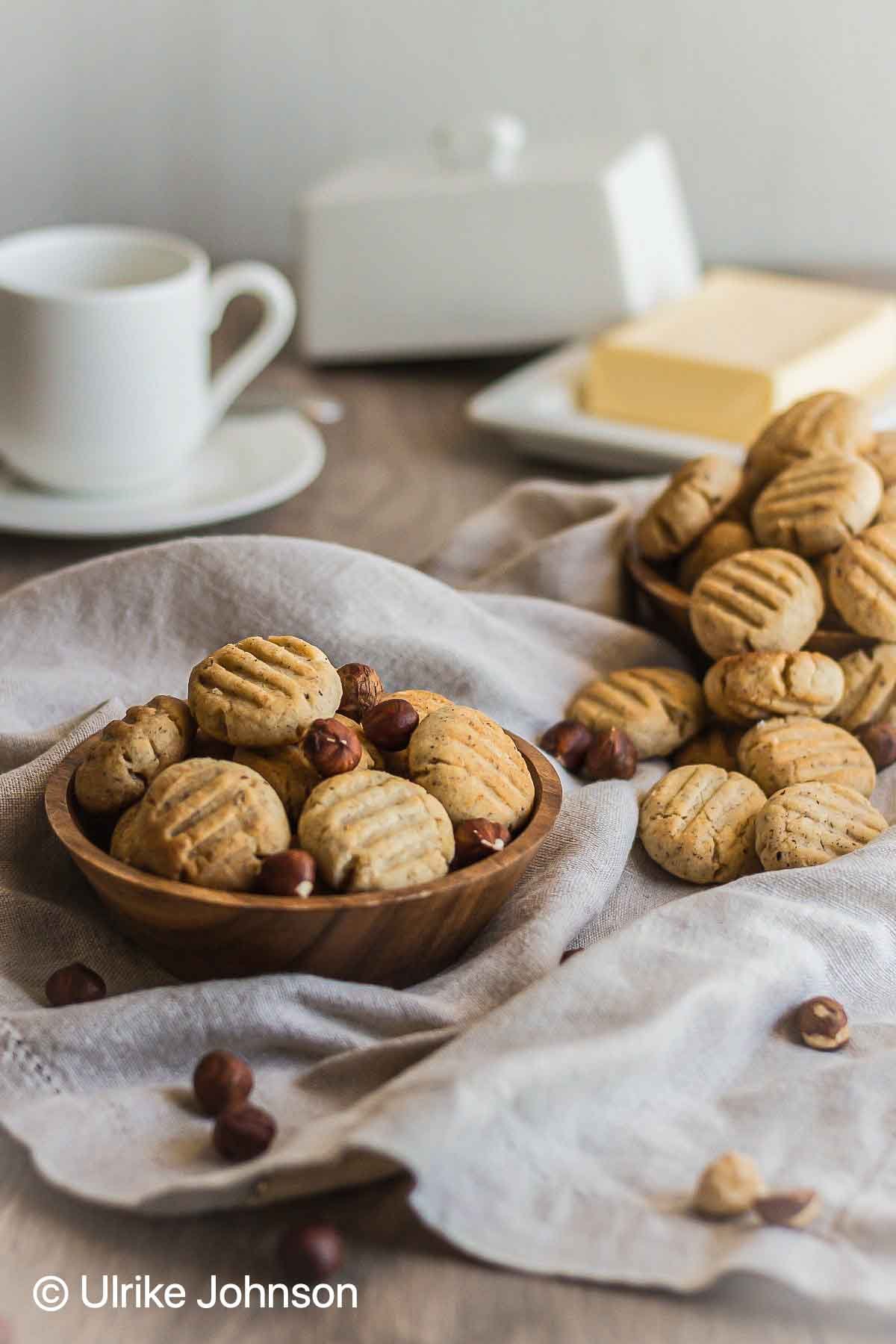 German hazelnut cookies in a wooden bowl surrounded with whole hazelnuts and a butter dish in the background