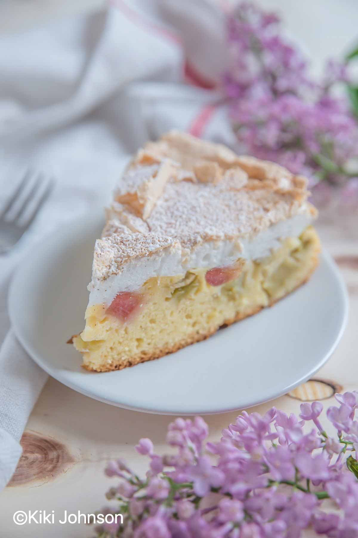 a slice of German Rhubarb Meringue Cake on a white cake plate with some lilac flowers in the background