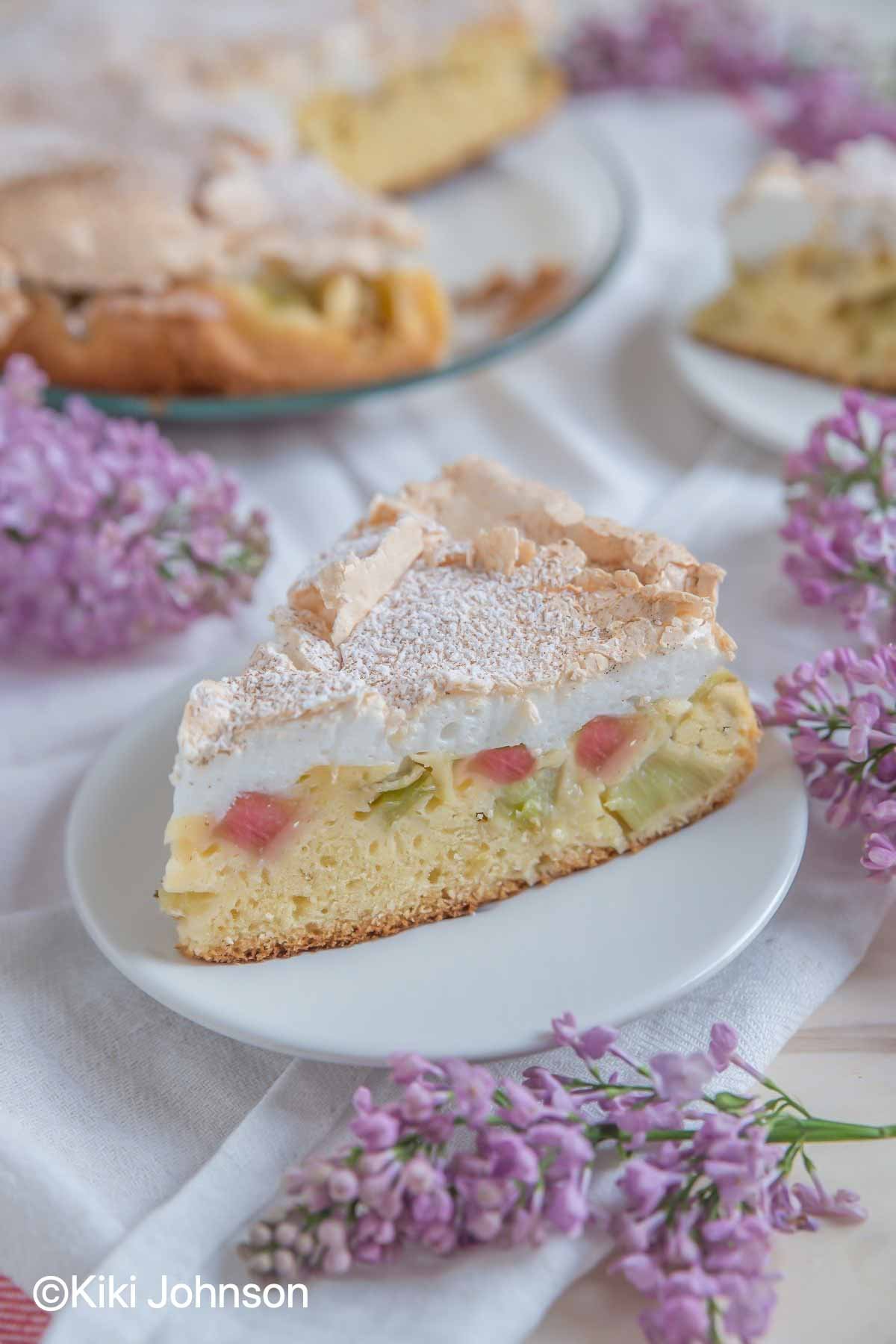 a slice of German Rhubarb Meringue Cake on a white cake plate surrounded with lilac flowers
