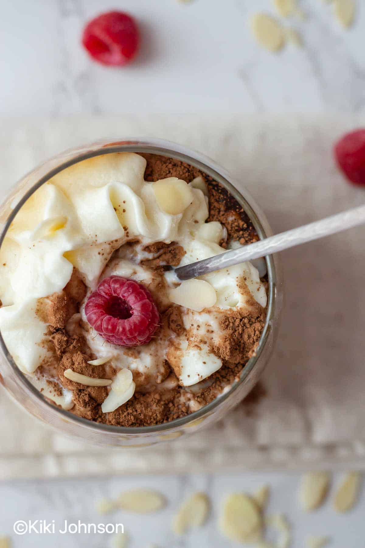 a spoon being dipped in a mason jar with tiramisu overnight oats decorated with raspberries and cocoa powder.