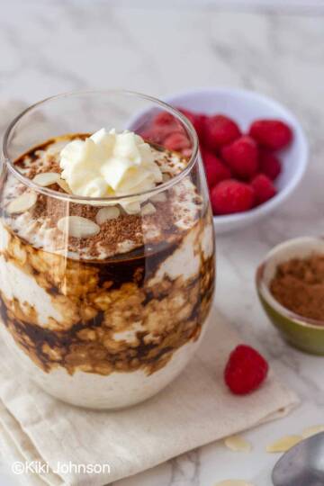 tiramisu overnight oats topped with cocoa powder in a glass with fresh raspberries in the background