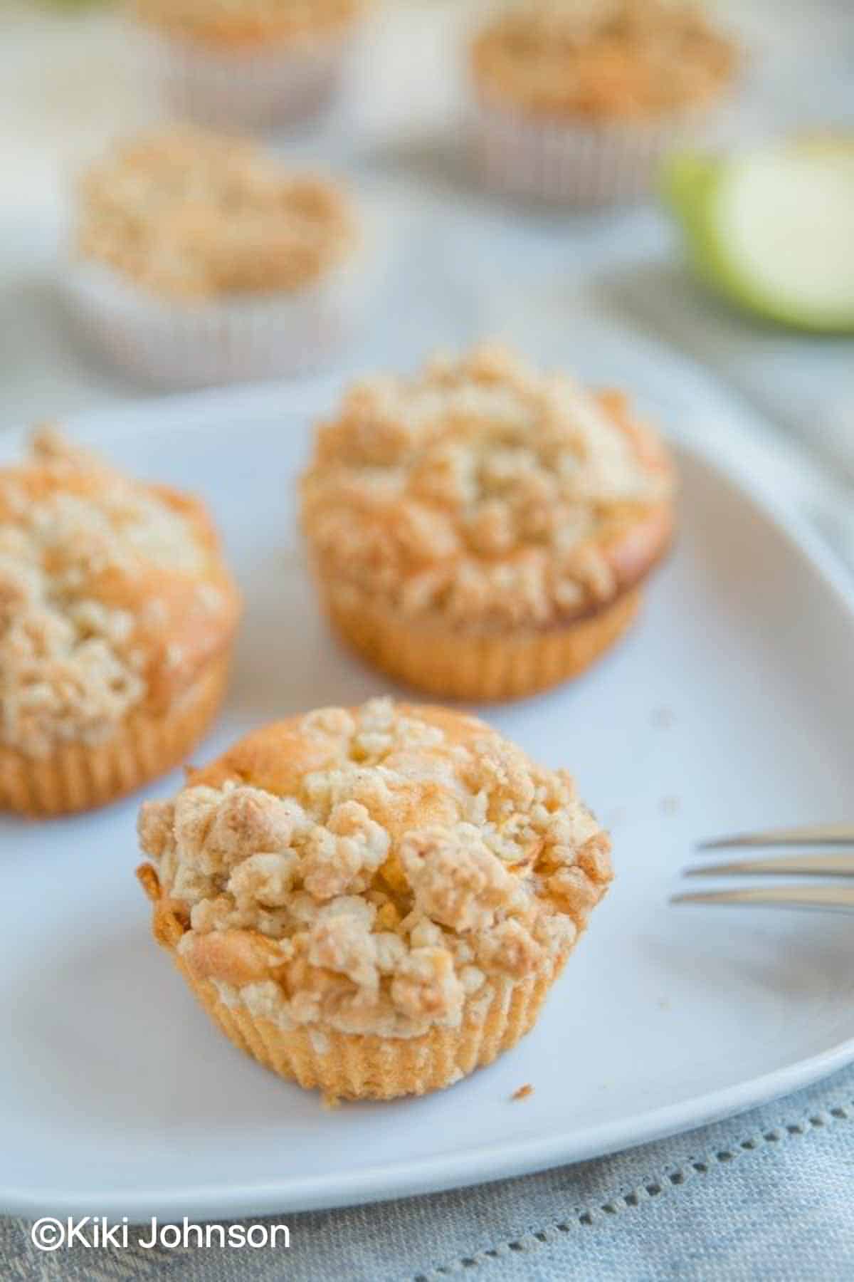 three German Apple Muffins with Crumb topping on a white plate with a fork on the side 