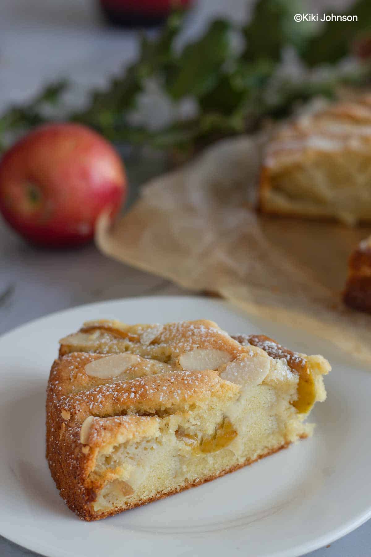 a slice of German apple almond cake on a dessert plate with fresh apples in the background
