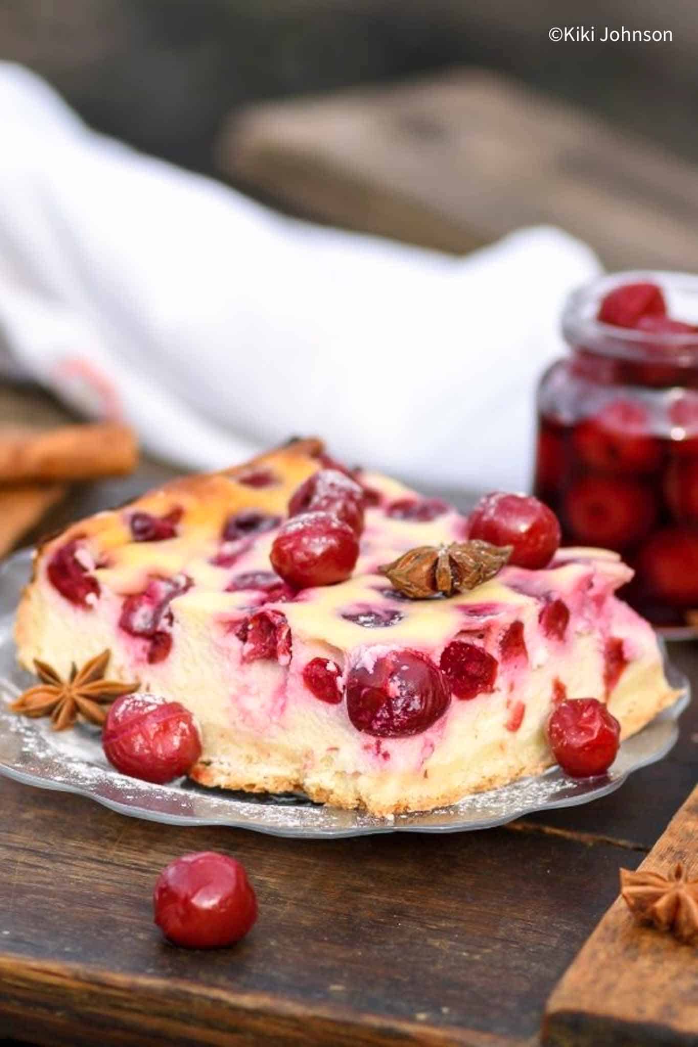 a slice of Oma's German cherry cheesecake topped with cherries and cloves with a jar of canned sour cherries in the back