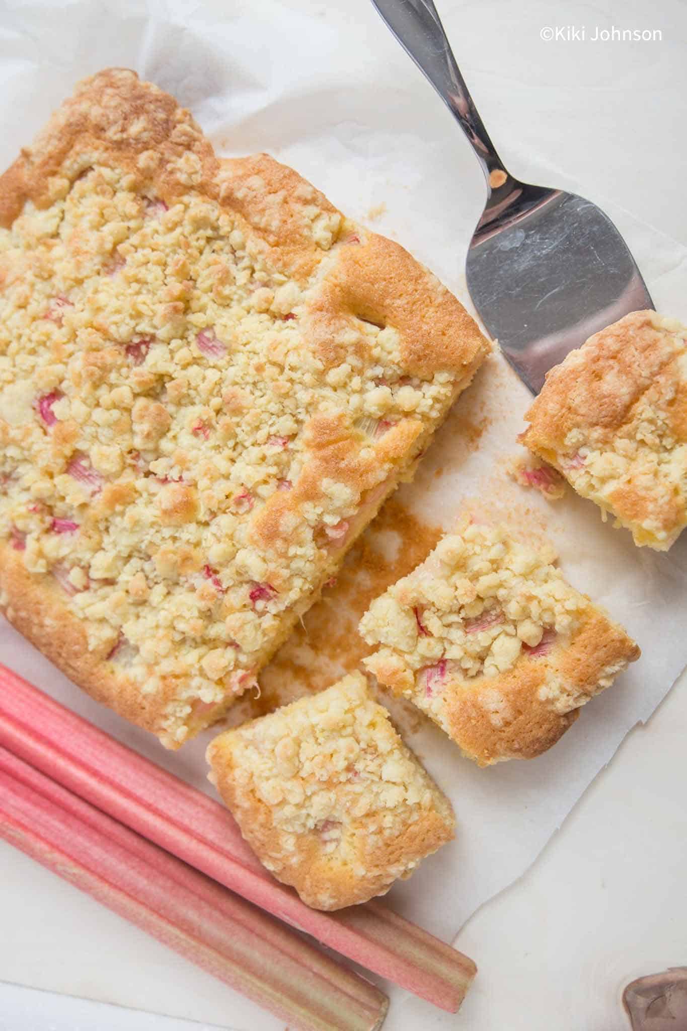 Rhabarber Blechkuchen mit Marzipanstreusel auf einem Stück Backpapier neben frischen Rhabarberstangen