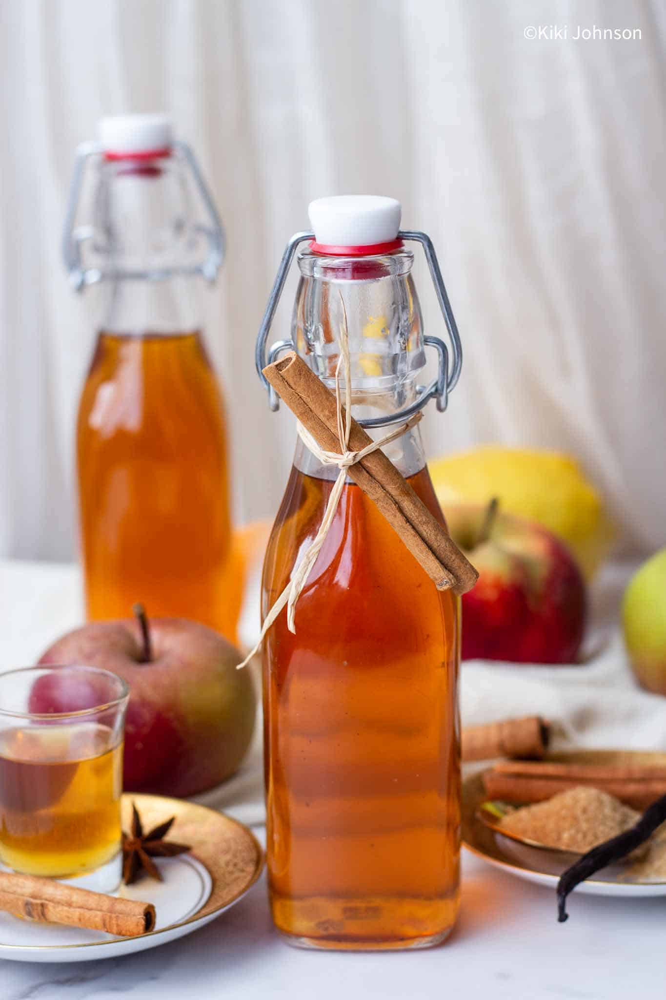 a bottle of homemade apple liqueur next to cinnamon sticks and fresh apples.