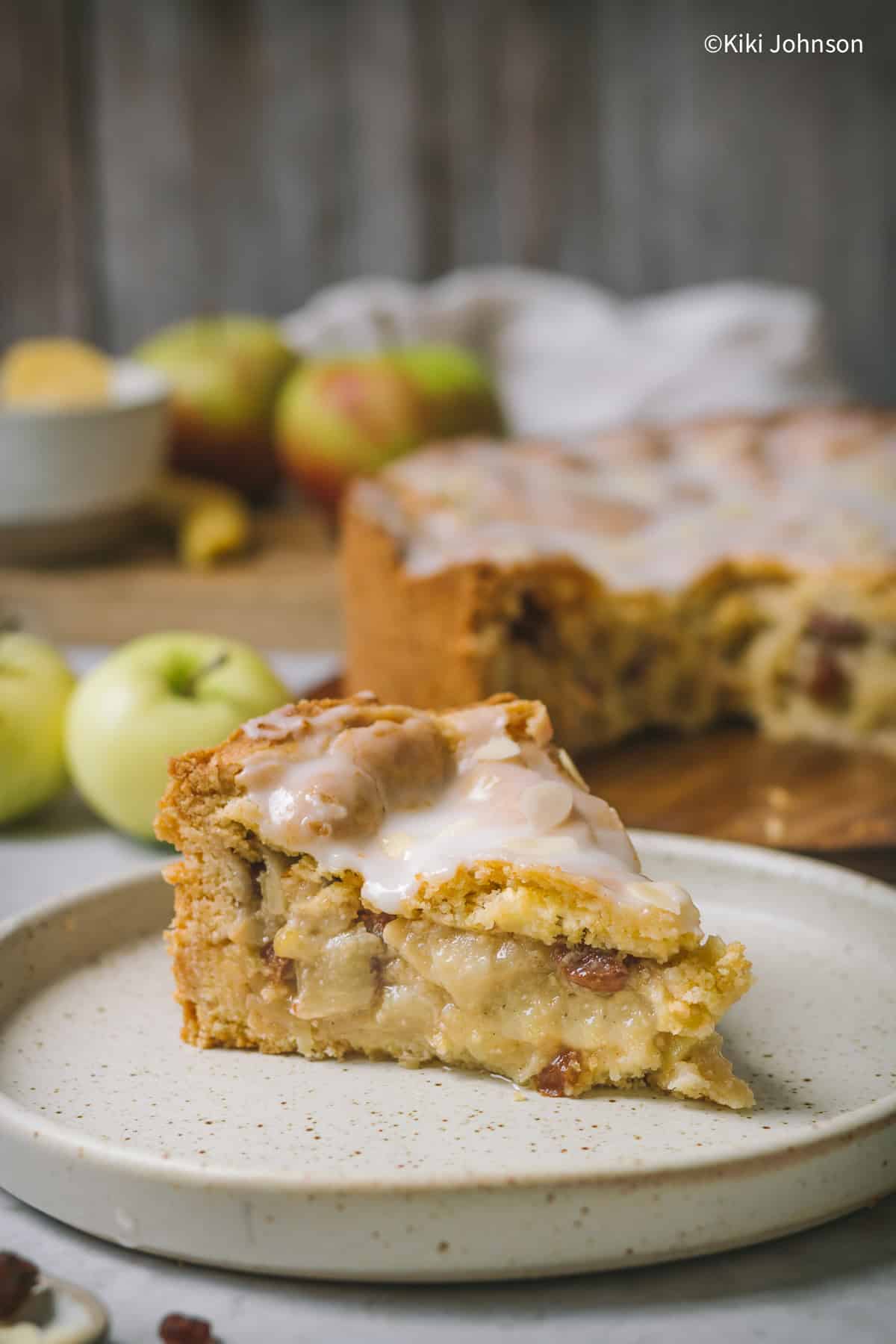 ein Stück  Landfrauen Apfelkuchen mit Rumrosinen und Zuckerguss mit einem angeschnittenen Apfelkuchen im Hintergrund. 