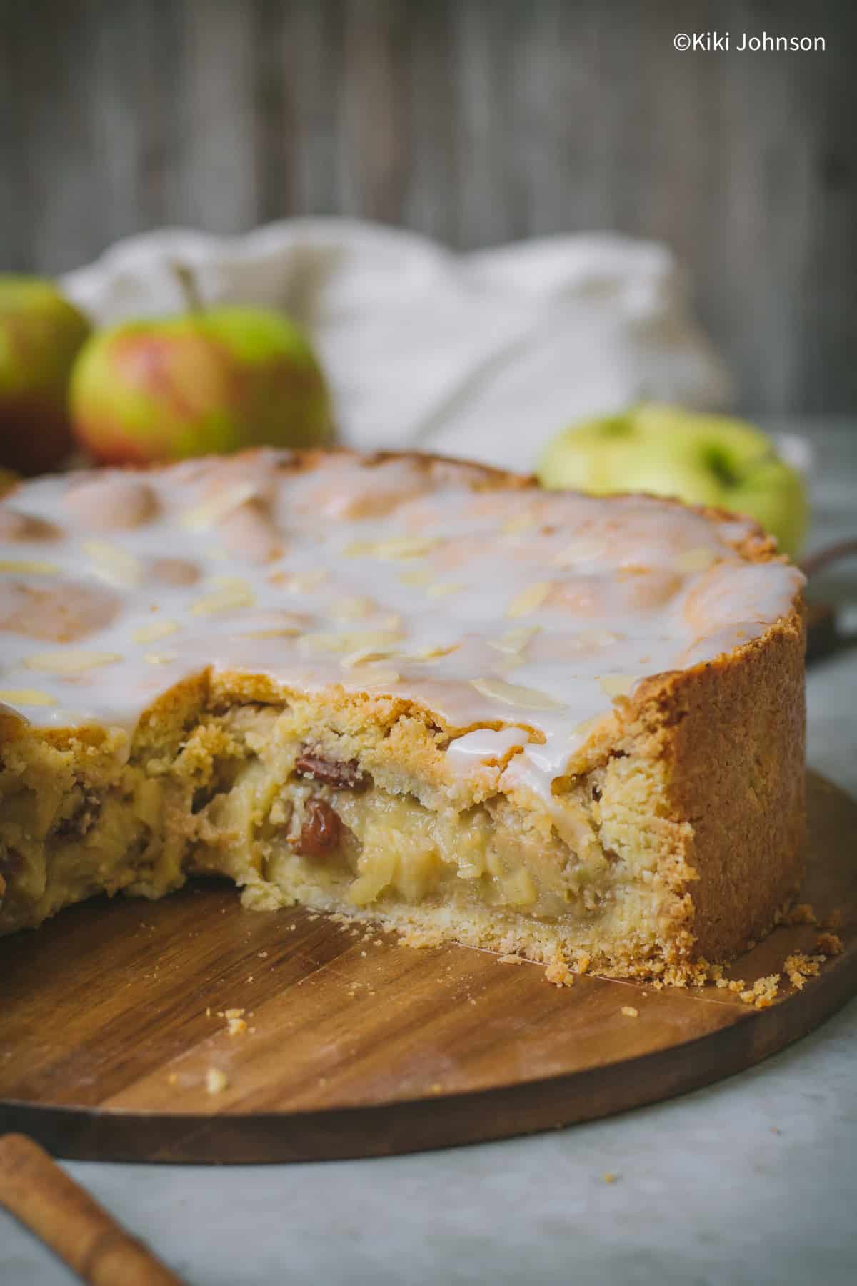 a German Apple Pie on a wooden board filled with apples, raisins and lemon glaze with 1 piece being cut out