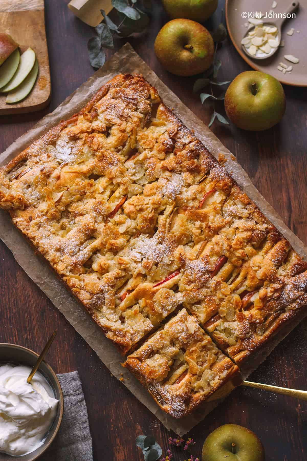 ein angeschnittener Apfelkuchen vom Blech mit Mandeln und Puderzucker dekoriert neben einer Schale mit Schlagsahne