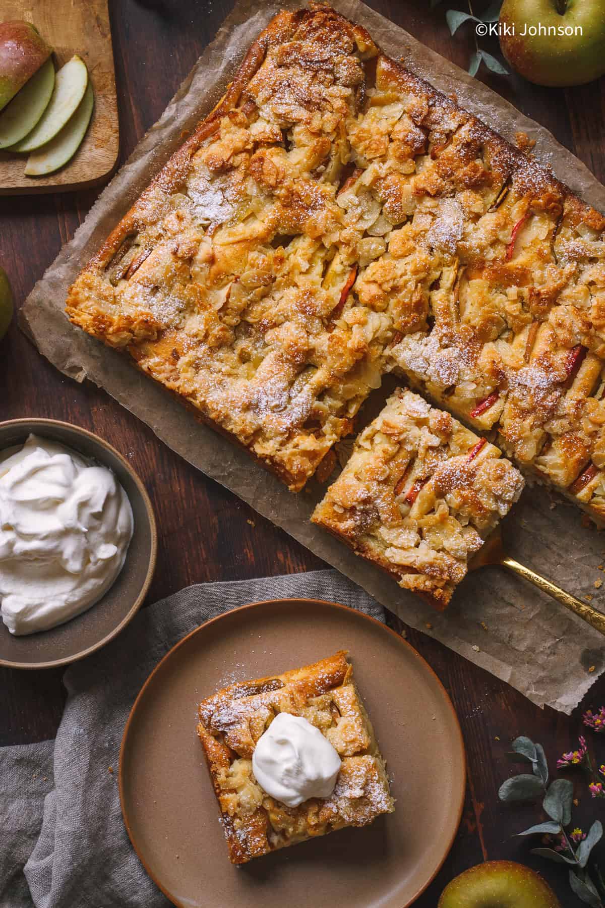 ein Stück Landfrauen Apfelkuchen vom Blech mit einem Klecks Sahne serviert neben einem Backblech mit mehr Kuchen.