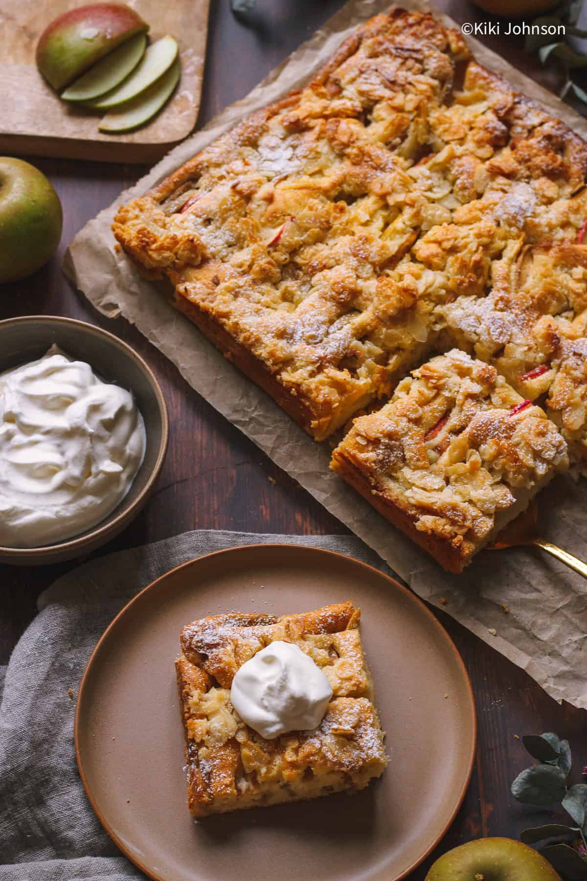 traditional German apple sheet cake on a sheetpan with one piece served on a plate with whipping cream.