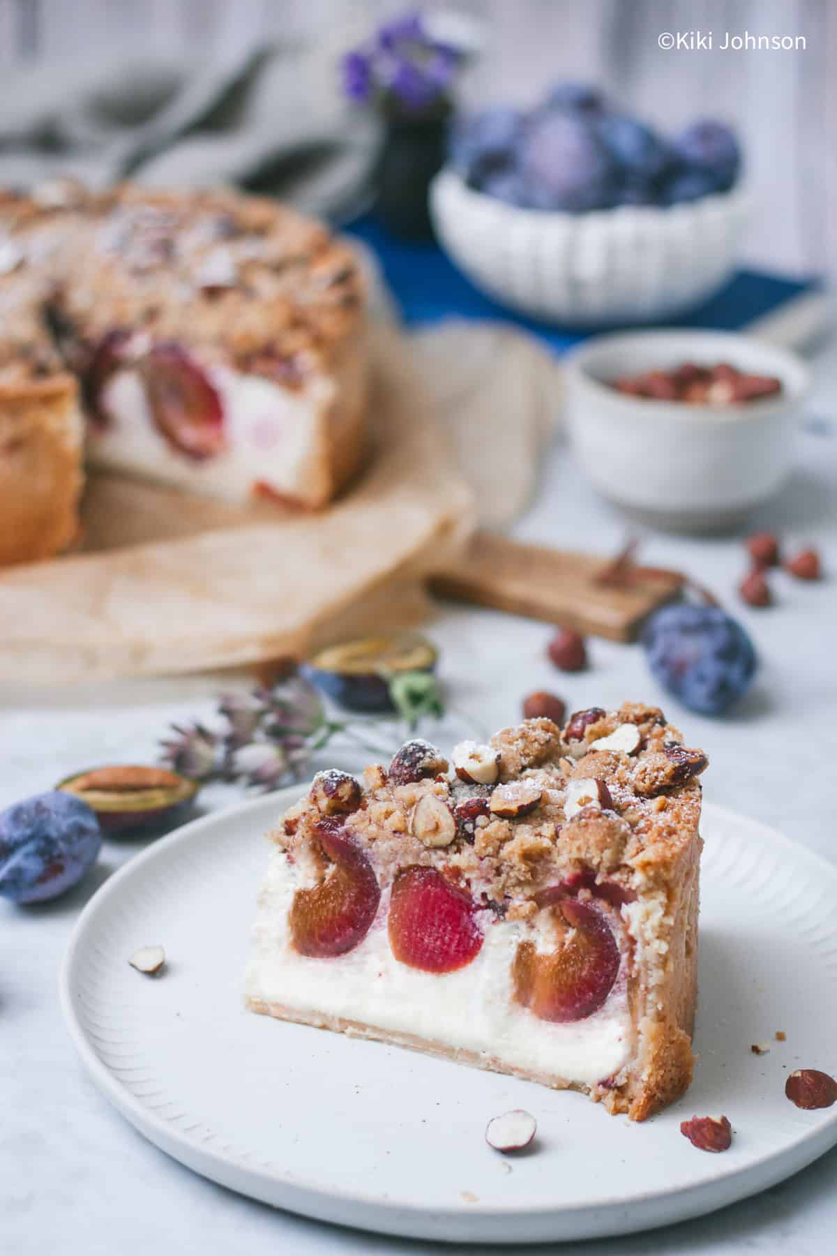 ein Stück Käsekuchen mit Pflaumen und Nuss Zimtstreuseln mit frischen Zwetschgen im Hintergrund.