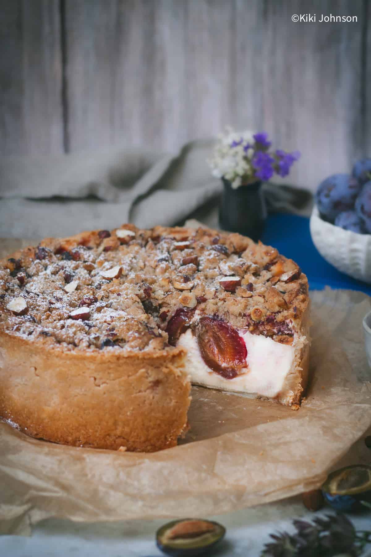 ein angeschnittener Pflaumen Käsekuchen mit Streuseln und Haselnüssen auf einem Holzbrett.