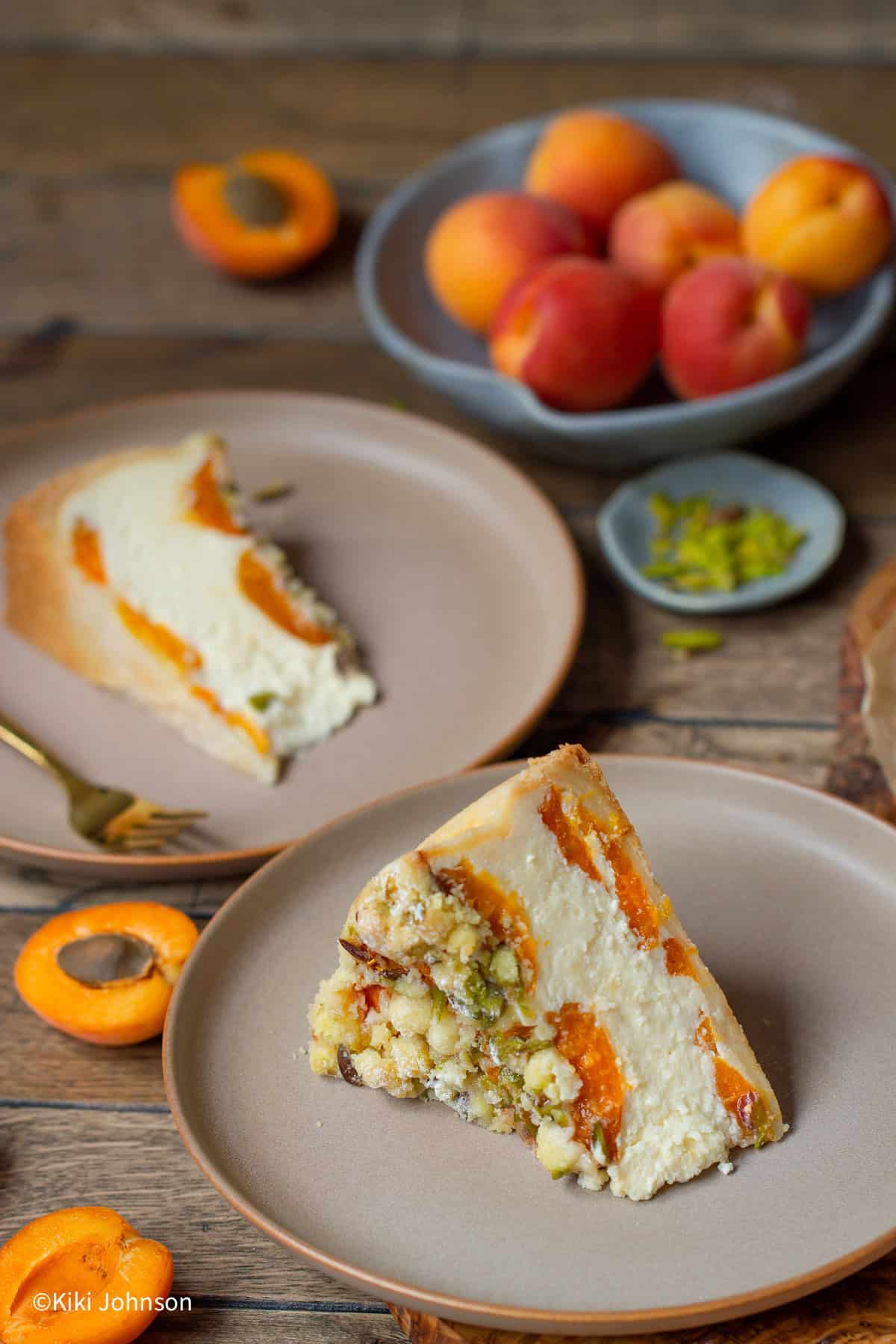 zwei Stücke Marillenkuchen mit Topfen und Pistazien Streusel auf zwei braunen Tellern mit frischen Aprikosen im Hintergrund