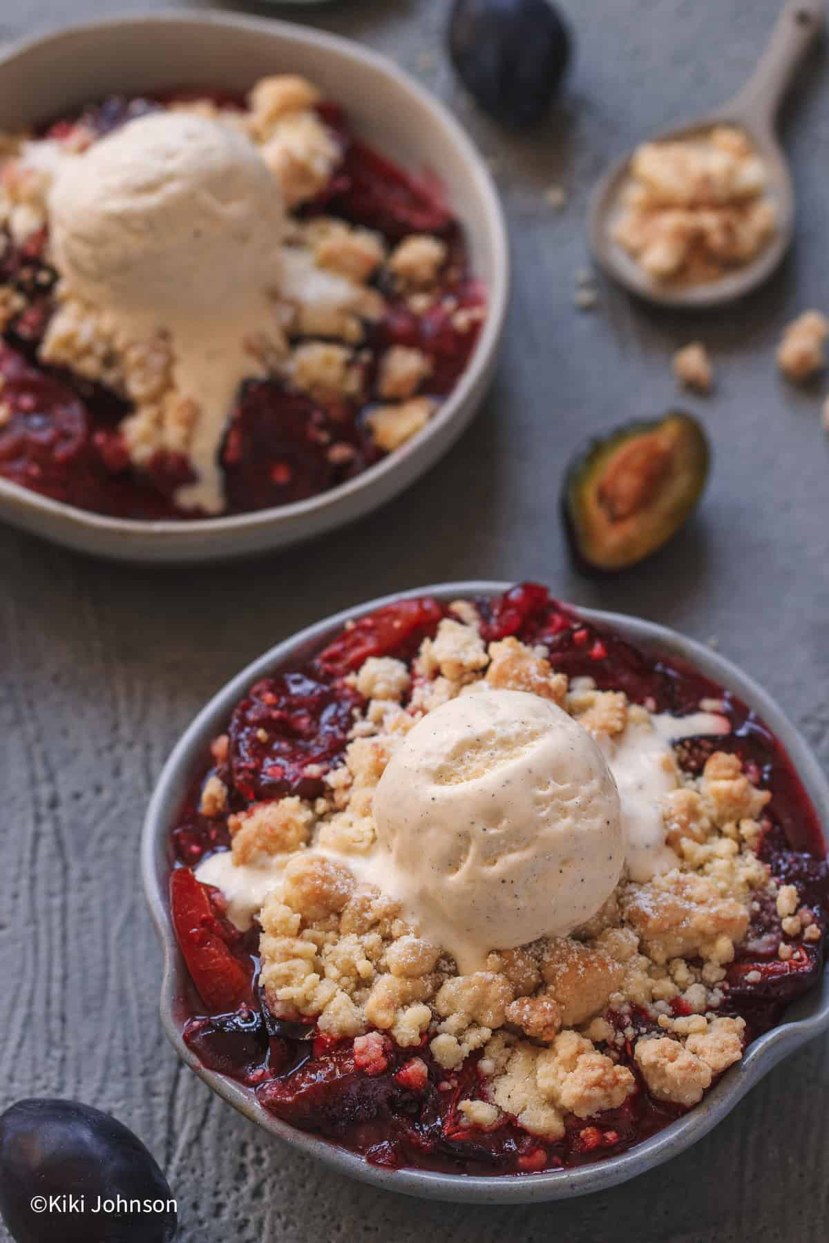 NAhaufnahme von einer Schale zwetschgen Crumble mit einer halbgeschmolzenen Kugel Vanilleeis mit ein paar aufgeschnittenen Zwetschgen daneben