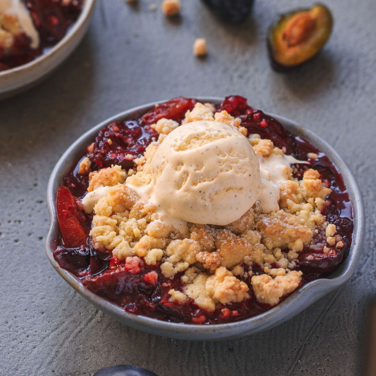 eine Portion Zwetschgen Crumble serviert mit einer Kugel Vanilleeis in einer kleinen blauen Dessertschale