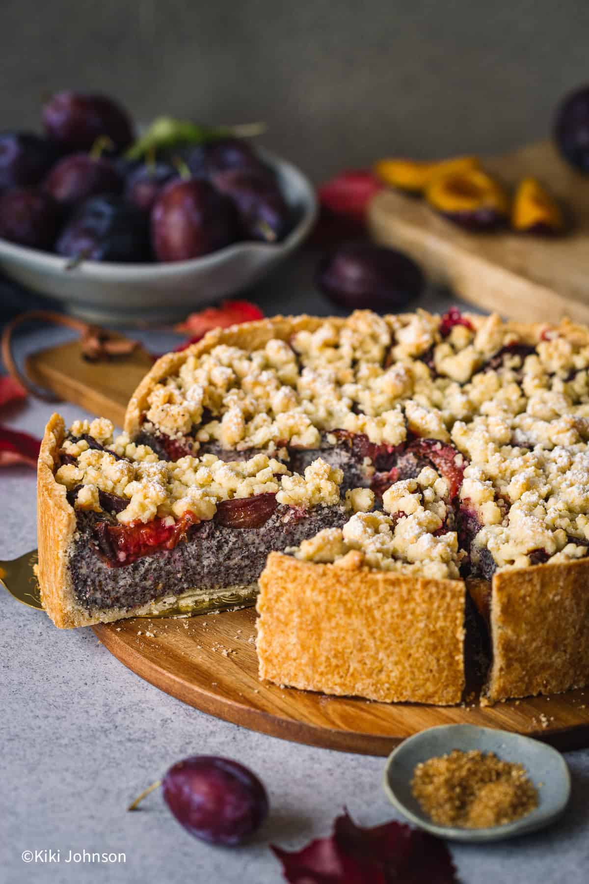 Ein Saftiger Zwetschgen Mohnkuchen mit Pudding und Streuseln auf einer Holz Kuchenplatte mit frischen Zwetschgen im Hintergrund.