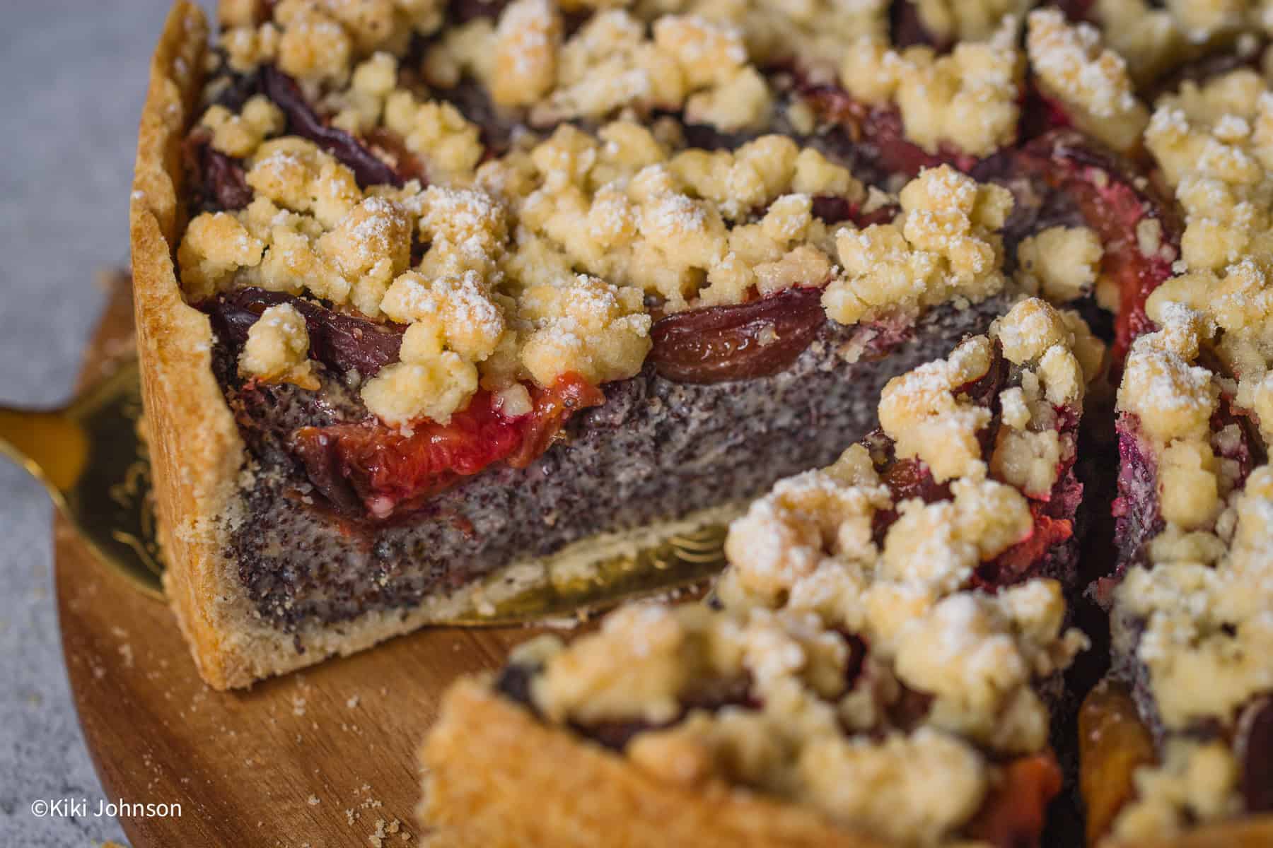 ein Kuchenheber hebt ein Stück saftigen Mohnkuchen mit zwetschgen und Streuseln von einem Holzbrett.