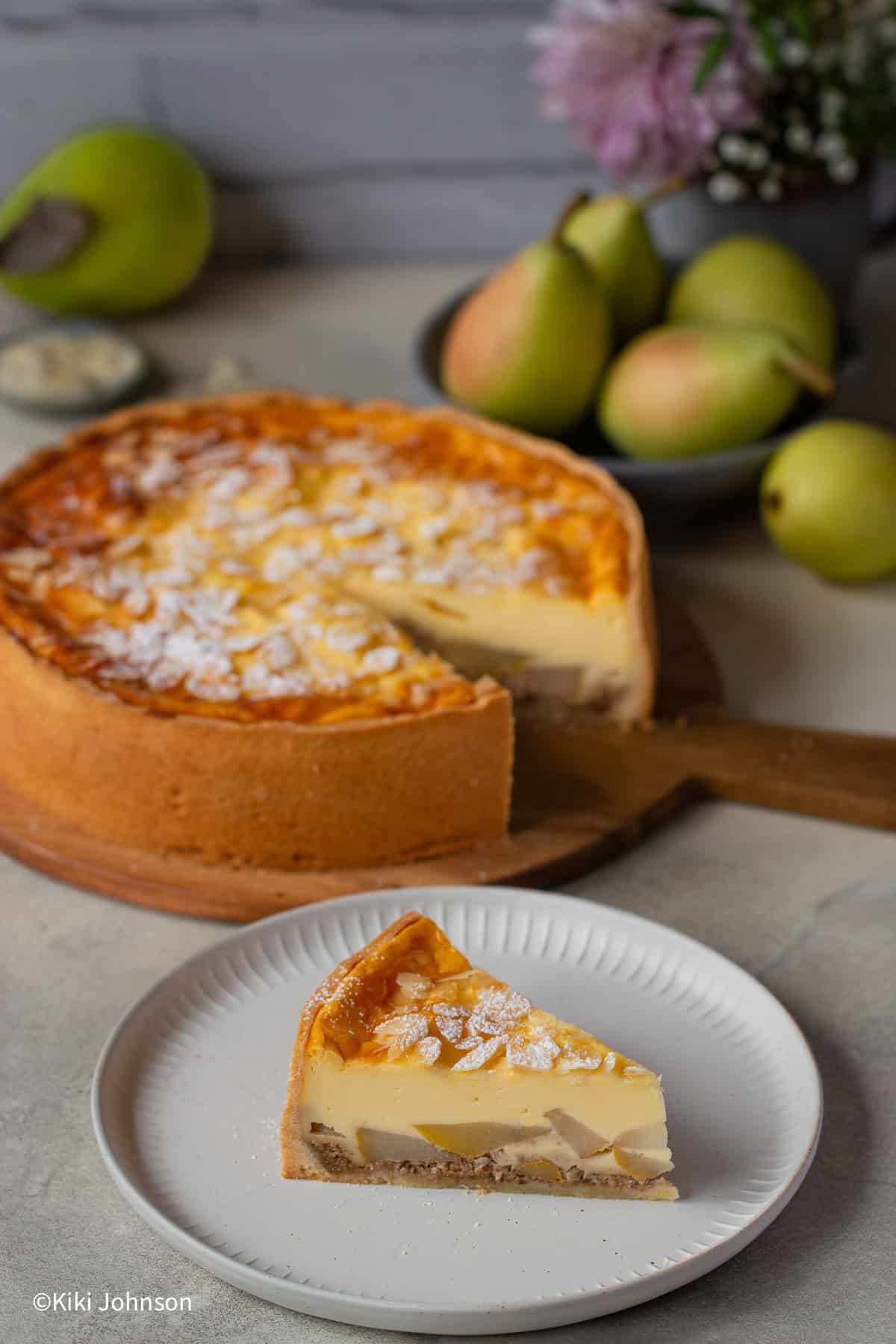 ein Stück Birnenkuchen mit Pudding Schmand GUss und Walnüssen mit einem angeschnittenen Kuchen und Herbstblumen im Hintergrund