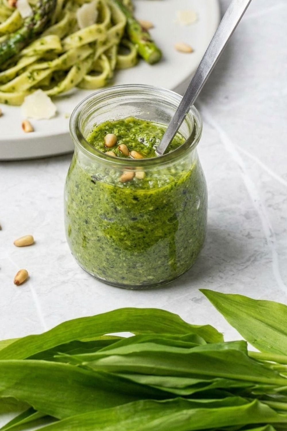ein Glas Bärlauch Pesto mit einem Teller Pasta mit Spargel und Bärlauch Pesto im Hintergrund