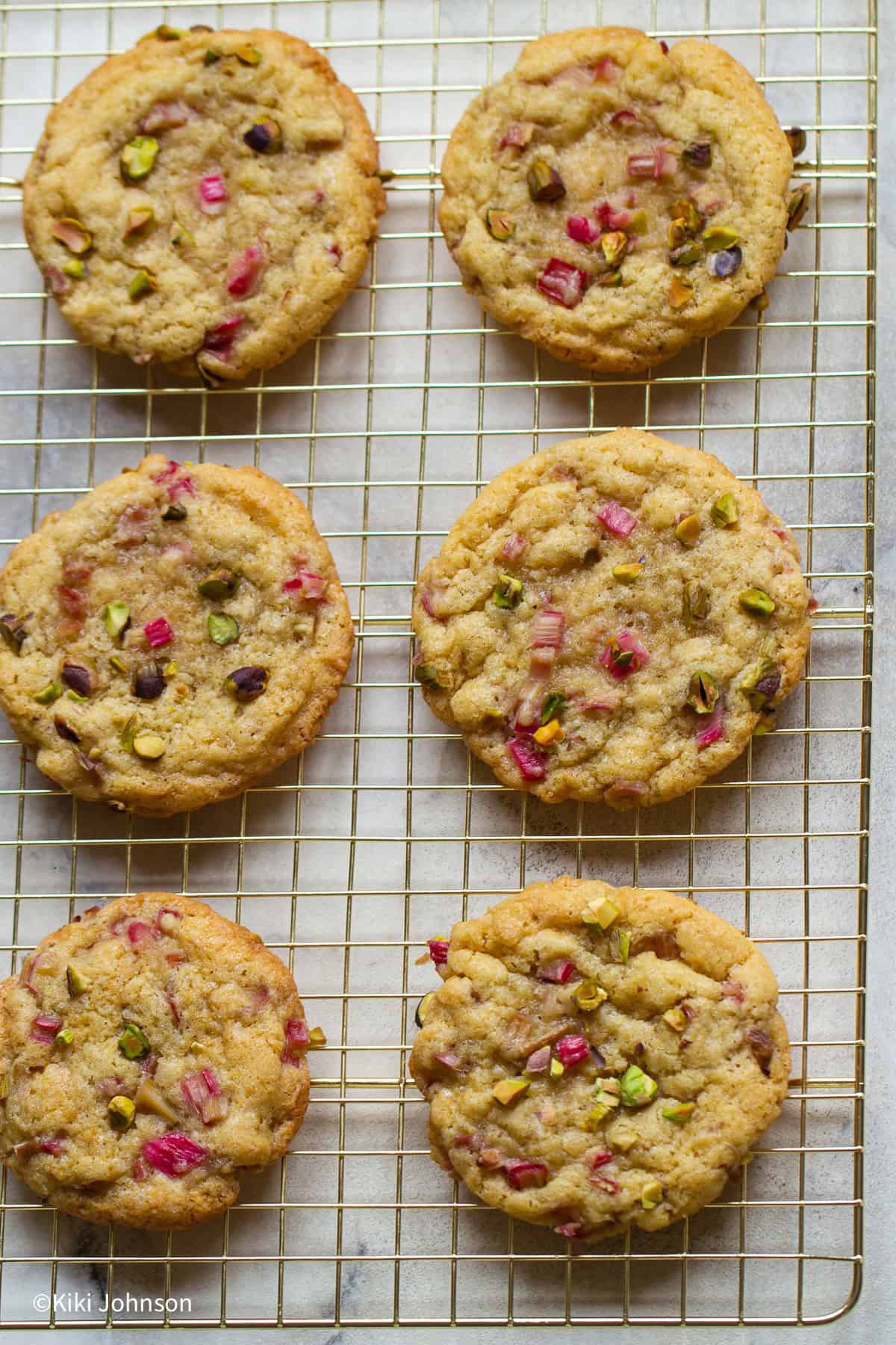 Chewy rhubarb pistachio cookies cooling on a wire rack, studded with pink rhubarb pieces and green pistachios, golden edges with soft centers.
