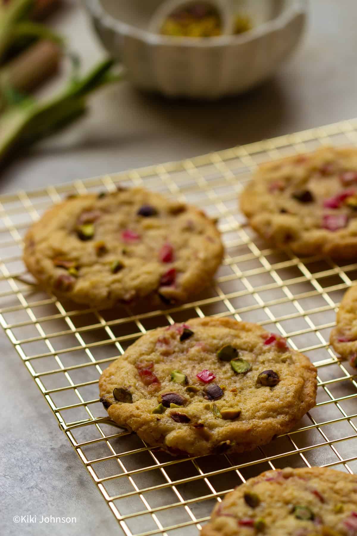 Rhabarber Pistazien Kekse auf einem goldenen Gitter mit frischen Rhabarber Stangen im Hintergrund 