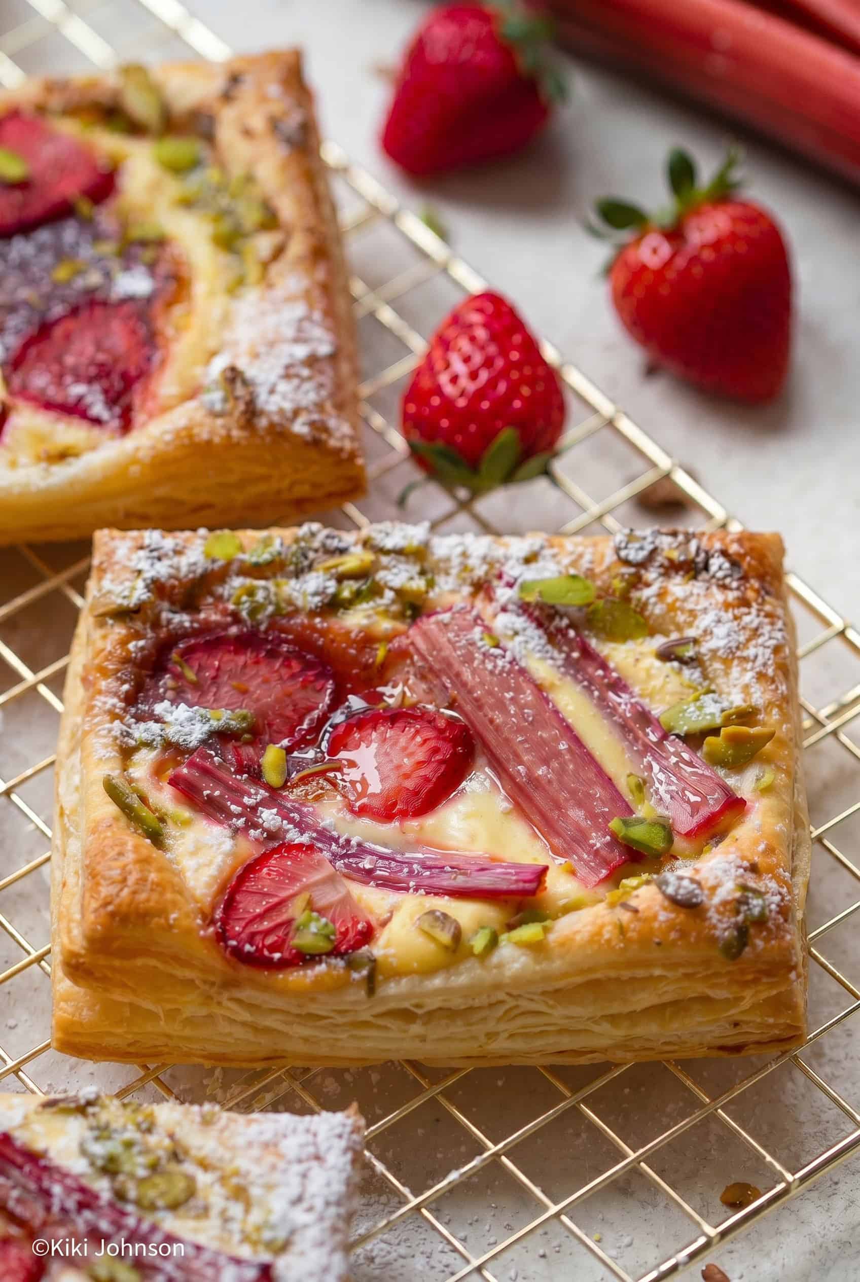Nahaufnahme von einem  Erdbeer Rhabarber Pudding Blätterteig Teilchen mit Quark Pudding Füllung auf einem goldenen Gitter  mit frischen Erdbeeren im Hintergrund 