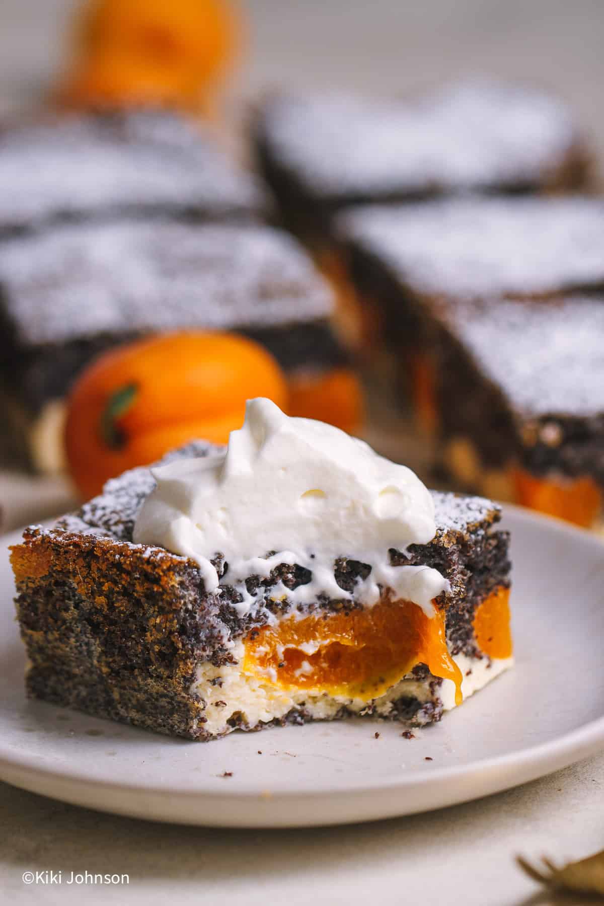 Ein halb aufgegessenes Stück Saftiger Aprikosenkuchen mit Mohn und Quark auf einem Teller mit Puderzucker bestäubt und mit einem Klecks Sahne serviert. Im Anschnitt sieht man die cremige Quarkschicht und die gebackenen Aprikosen im dunklen Mohnteig.