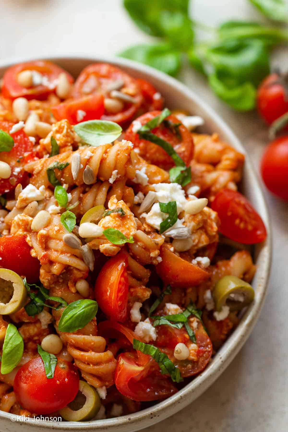 Nahaufnahme von Nudelsalat mit Pesto, Feta, Tomaten und Oliven in einer weissen Schale mit Sonnenblumenkernen dekoriert, daneben frischer Basilikum
