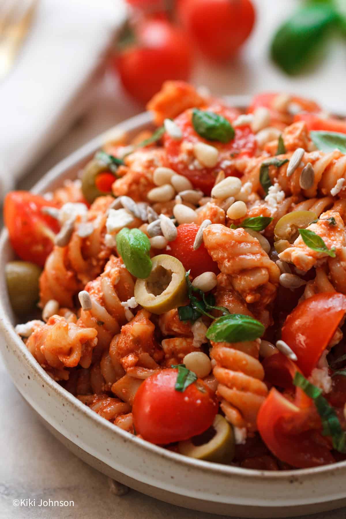 Nahaufnahme von Nudelsalat mit Pesto, Feta, Tomaten und Oliven in einer weissen Schale mit Sonnenblumenkernen dekoriert, daneben frischer Basilikum