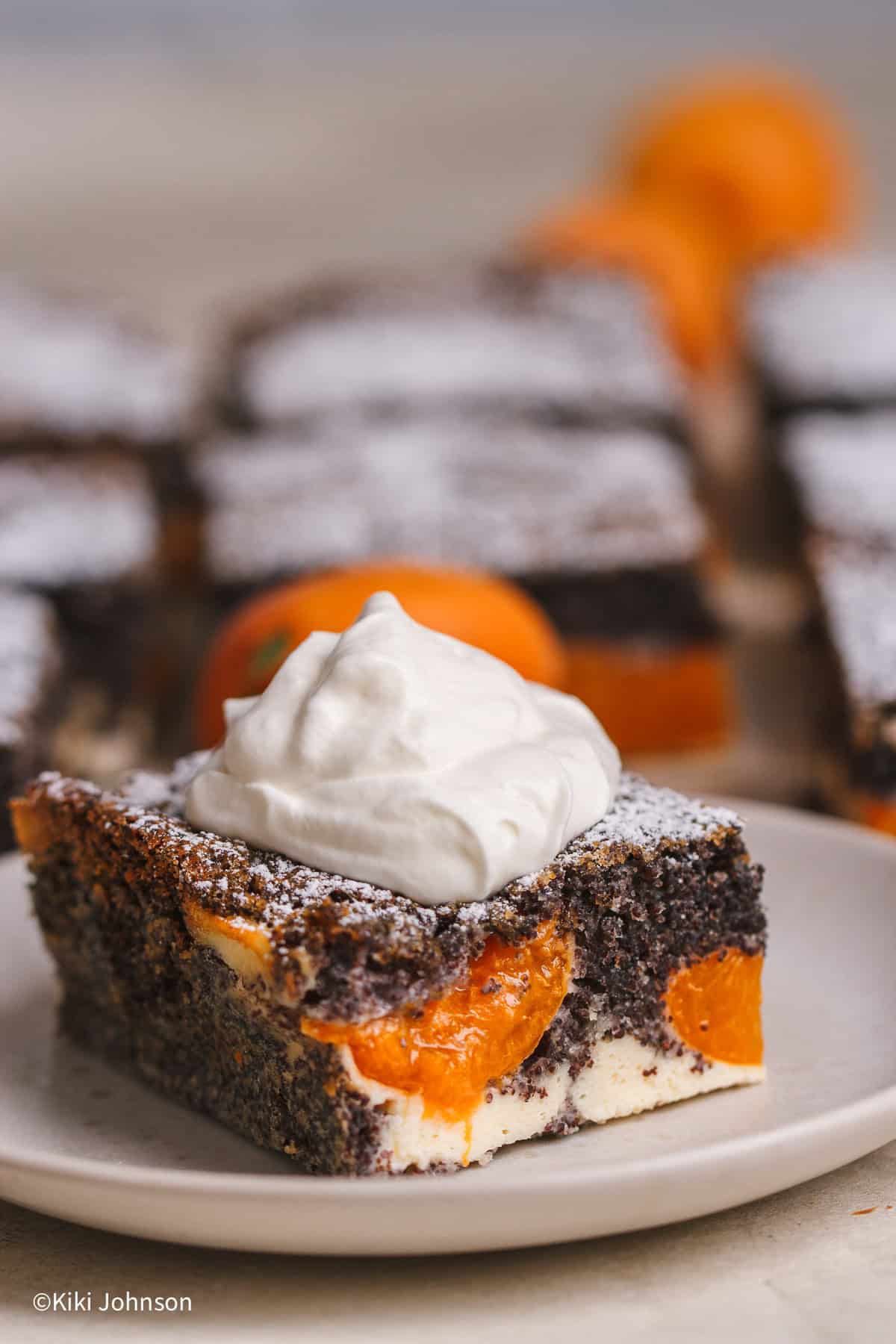 Ein Stück Saftiger Aprikosenkuchen mit Mohn und Quark auf einem Teller mit Puderzucker bestäubt und mit einem Klecks Sahne serviert. Im Anschnitt sieht man die cremige Quarkschicht und die gebackenen Aprikosen im dunklen Mohnteig.