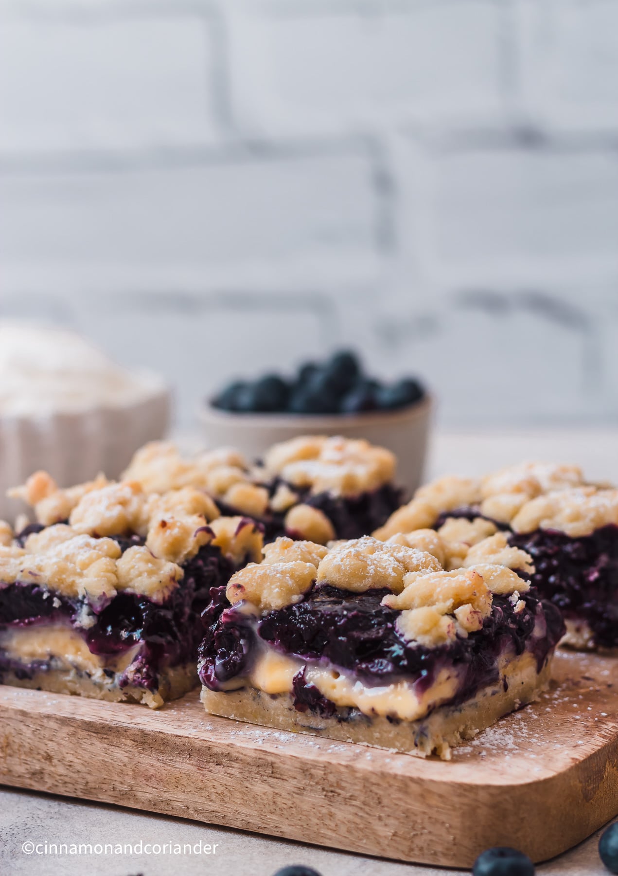 vier Stück Blaubeerkuchen mit Pudding und Streuseln vom Blech mit einer schale frischen Blaubeeren und Sahne im Hintergrund