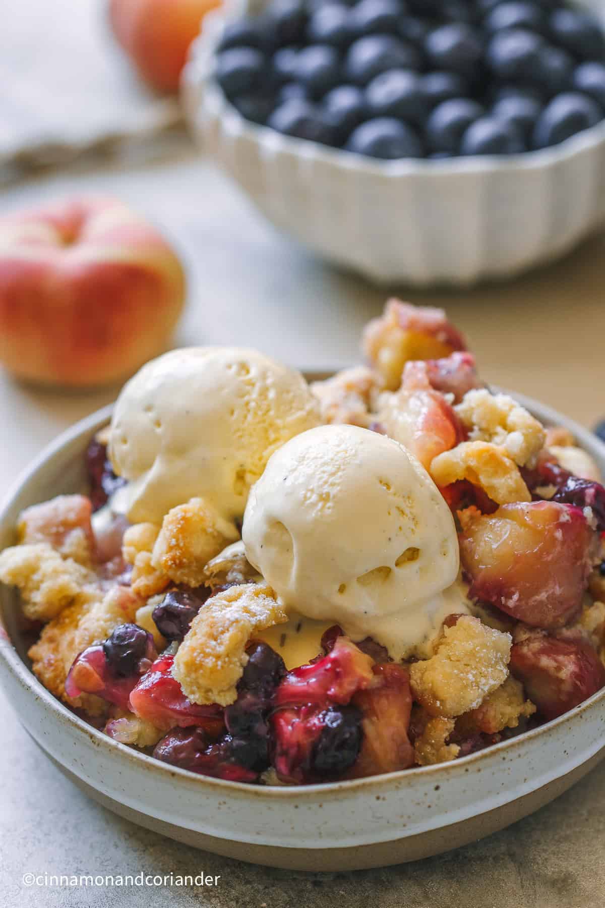 eine Schale mit Pfirsich Blaubeer Crumble mit 2 Kugeln Vanille Eis serviert mit WEinbergpfrisichen und frischen Blaubeeren  im Hintergrund