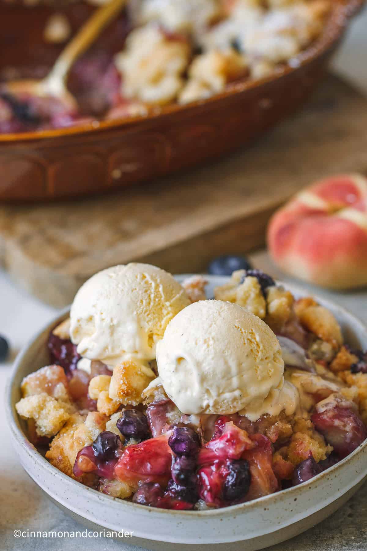eine Schale mit Pfirsich Blaubeer Crumble mit 2 Kugeln Vanille Eis serviert mit WEinbergpfrisichen im Hintergrund