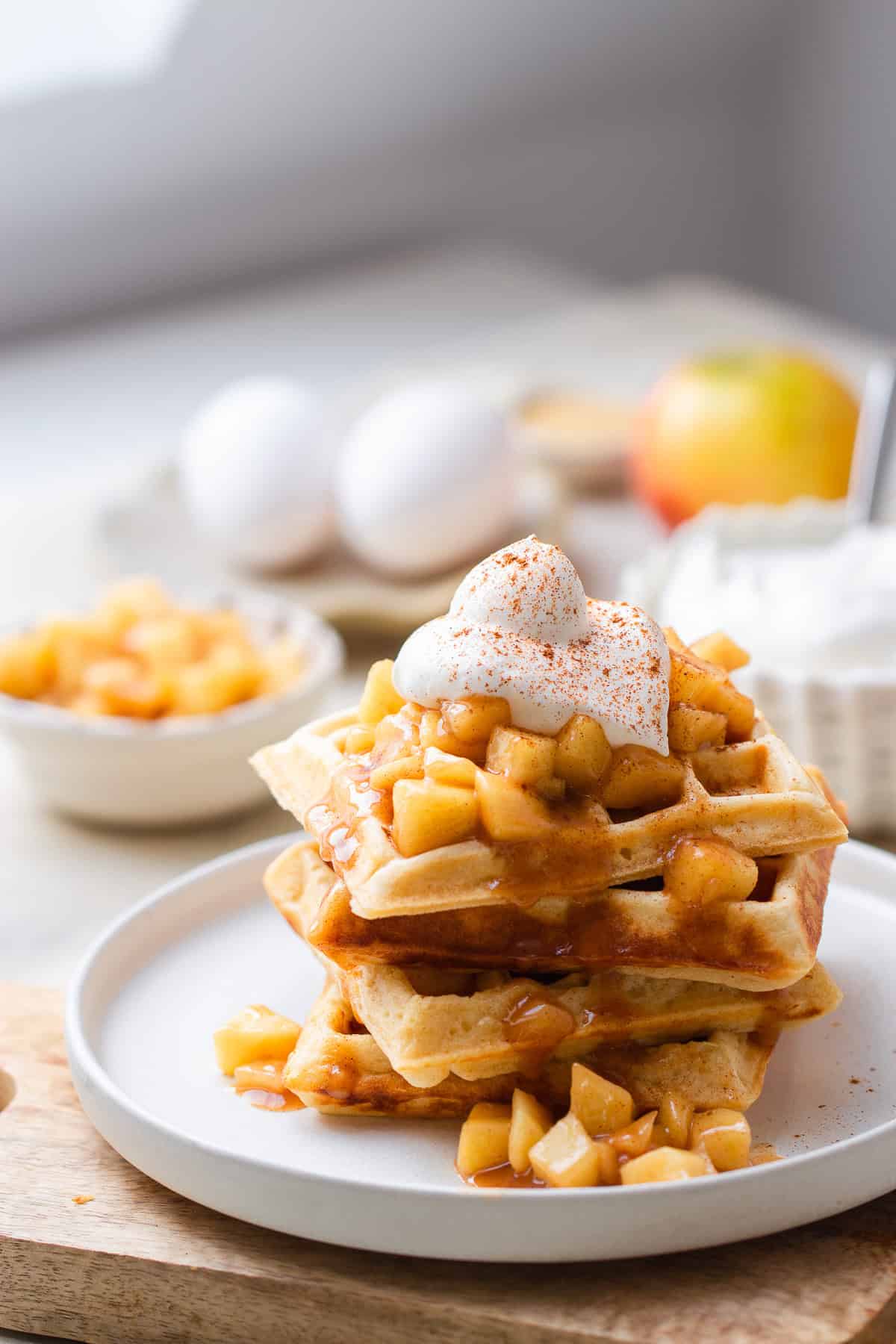 ein Stapel Hefewaffeln mit Apfel Zimt Topping und Sahne serviert mit einer SChale Apfelkompott im Hintergrund